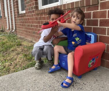 Two young boys hold a red styrofoam airplane in what appears to be a backyard. Cayden is seated in a small, red and blue "PJ Masks" chair, while LJ crouches next to him. The chair is on a cement slab against a brick house.