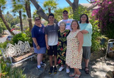 A group of six people, three men and three women, pose for a family photo on the Fourth of July. They're standing outside of an assisted living facility on a stone walkway leading to a bench. Lush green plants, palm trees, and bougainvillea surround them.