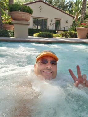 A man with an orange visor and sunglasses shows the peace sign as he's in the water, with hot tub bubbles around him. In the rear are the pool patio, low green shrubs and what may be a clubhouse.