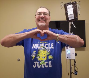 A man in a purple T-shirt that says "Muscle Juice" stands next to an IV pole and makes a heart shape with his hands. 