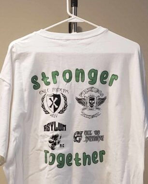 A white T-shirt with black and green lettering hangs on a plastic hook. The T-shirt says "Stronger Together."
