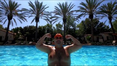 A man stands shirtless in a large swimming pool with palm trees in the background. He wears a swimming cap and flexes both of his biceps in the typical upper-body muscular pose.