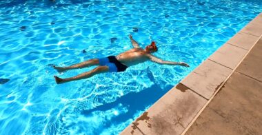 A photo from the perspective of someone standing by the side of a pool shows a man in swimming trunks floating, with arms stretched out to the side. 
