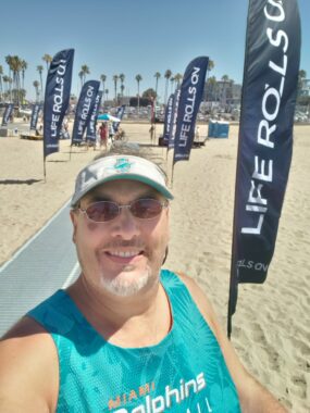 A man in a white Miami Dolphins visor and a green Miami Dolphins tank top stands on a beach, which has Life Rolls On banners on poles on either side of what looks like a gray runner. Palm trees are in the distance. The man has a goatee and mustache and wears sunglasses.