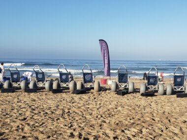 A line of wheelchairs with large wheels are on a beach facing the water, with a long, narrow purple banner in the middle.