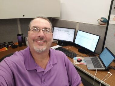 A goateed man wearing a purple polo shirt and glasses takes a selfie in his cubicle at work. A laptop, keyboard, and several monitors are visible on his desk in the background.