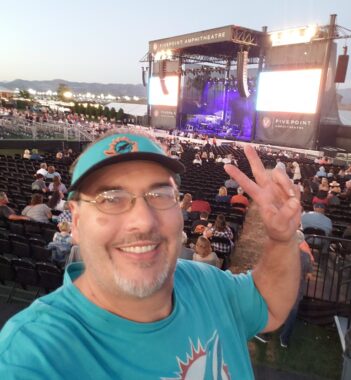 Outdoors, a man with glasses and a light goatee and mustache flashes the peace sign in the bleachers, with a concert stage at a distance. He wears a Miami Dolphins green sun visor and T-shirt.