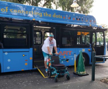 A blurry photo shows a man dressed in a white T-shirt and aqua blue cap and shorts - the colors of the Miami Dolphins - descending from a public bus on a ramp. He's pushing an aqua blue small wheelchair.