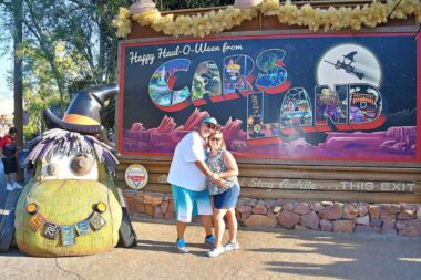 A husband and wife hug each other and pose for a photo in front of a sign at Disney California Adventure Park. It's hard to read the sign behind them, but it partially reads "Cars Land."
