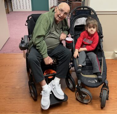A frail man in his 70s, bald and wearing glasses, sits in a motorized wheelchair next to a young boy who sits in his own mobility aid in what appears to be a home. The two are making happy, funny faces. The boy is wearing a red sweatshirt and the man a green hoodie. The man's very white shoes and socks stand out against his black sweat pants and the two black wheelchairs. 