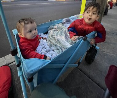 Two brothers, ages 5 years old and 10 months old, sit in a blue wagon on a sidewalk. The older boy is wearing a Spiderman costume, while the younger boy wears a red Incredibles costume. Both have blankets over their laps as they prepare to watch a Halloween parade.