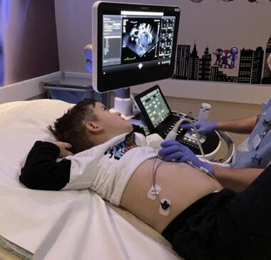 A young boy lies on his back on a hospital bed while a technician performs an echocardiogram. The boy's shirt is pulled up to his chest, and monitors are attached to his abdomen. The technician guides the ultrasound monitor over the boy's heart while the boy looks at the image on the screen.