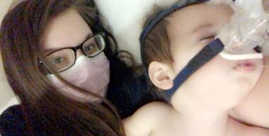 A woman with long brown hair, glasses, and a pink face mask lies next to an infant wearing a BiPAP mask over his nose.