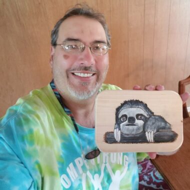A man with glasses and wearing a tie-dye T-shirt colored in light blue and green smiles while holding up a small wooden plaque with a sloth on it. 