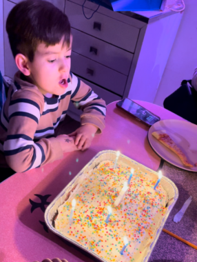 A young boy in a long-sleeved, striped shirt and with short, dark hair leans on a table and prepares to blow out six birthday candles on a yellow cake with candy sprinkles. 