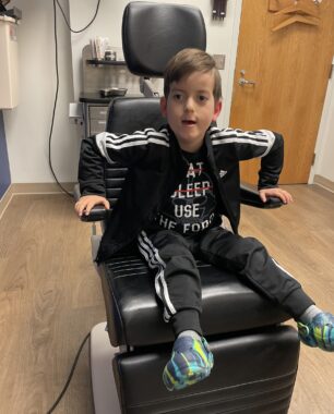 A 6-year-old boy sits in a chair in the office of an ear, nose, and throat specialist. He's wearing a sporty navy blue jumper and eagerly leans forward in the chair toward the camera, supporting himself with two hands on the arm rests of the chair. He looks at the camera with a coy expression.