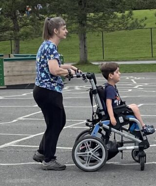A nurse walks behind a young boy in a wheelchair on a playground. Both are facing and headed to the right of the frame. A large park with grass and trees is visible in the background.