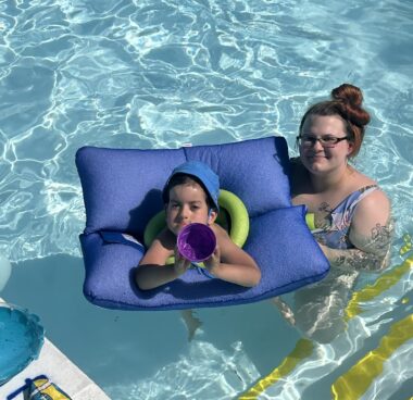 A young boy in a blue cap rests in a pool thanks to a square, blue float, with his head and arms poking through the middle. A woman with brown hair, glasses, and a multicolored bathing suit stands behind him in the water.