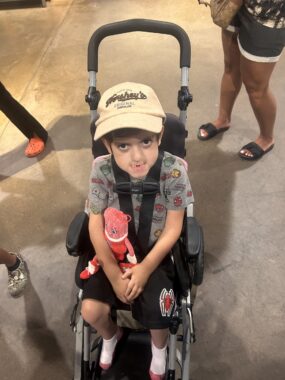 A young boy sits in a hybrid wheelchair/stroller and looks up at the camera. He is showing off a new Hershey's chocolate hat and a large "stuffed" Twizzler candy doll. 