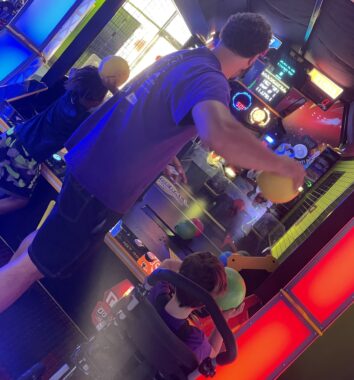 A young man gets ready to fire a yellow ball at a dodgeball video arcade game. The photo is taken from behind him, and to his left and right are two young boys, the one on the right is in a wheelchair. 