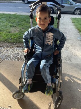 A boy in the first grade sits in his wheel chair in a blue and gray Nike jogging suit and hoodie. He is outside on the sidewalk, near a road. 