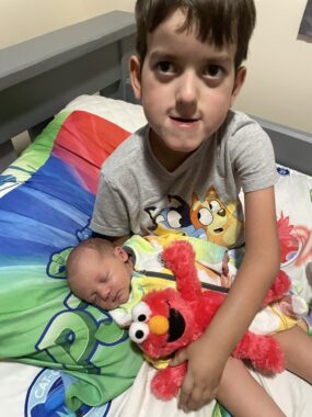 A boy looks up at the camera as he holds a newborn on his lap in a child's bed. In the same embrace is a red Elmo stuffed animal. The baby appears to be sleeping. 