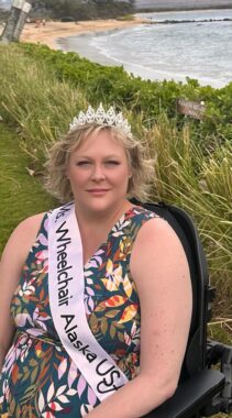 A woman with short blond hair poses for a photo near a beach. She's wearing a colorful patterned dress, a tiara, and a sash that reads "Ms. Wheelchair Alaska USA." She's seated in a black wheelchair with grass and green shrubs surrounding her. Water gently laps on a sandy beach in the background.