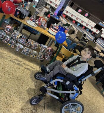 A boy in a lightweight wheelchair smiles for the camera in front of a Captain America-themed "Happy Birthday" sign at a bowling alley. The lanes are visible in the background. 