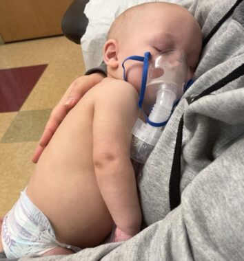 A baby rests with eyes closed rests on a lap, with someone's arms around him. He has a tube with a mask attached to his face to deliver a nebulizer treatment. 