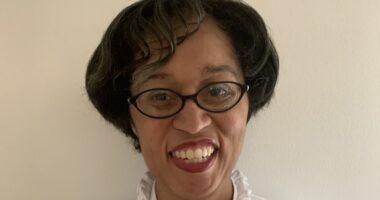 A woman, wearing glasses with short black hair, smiles at the camera.