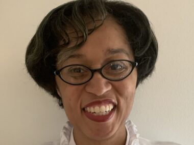 A woman, wearing glasses with short black hair, smiles at the camera.