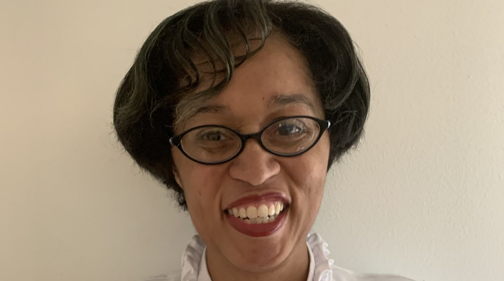 A woman, wearing glasses with short black hair, smiles at the camera.
